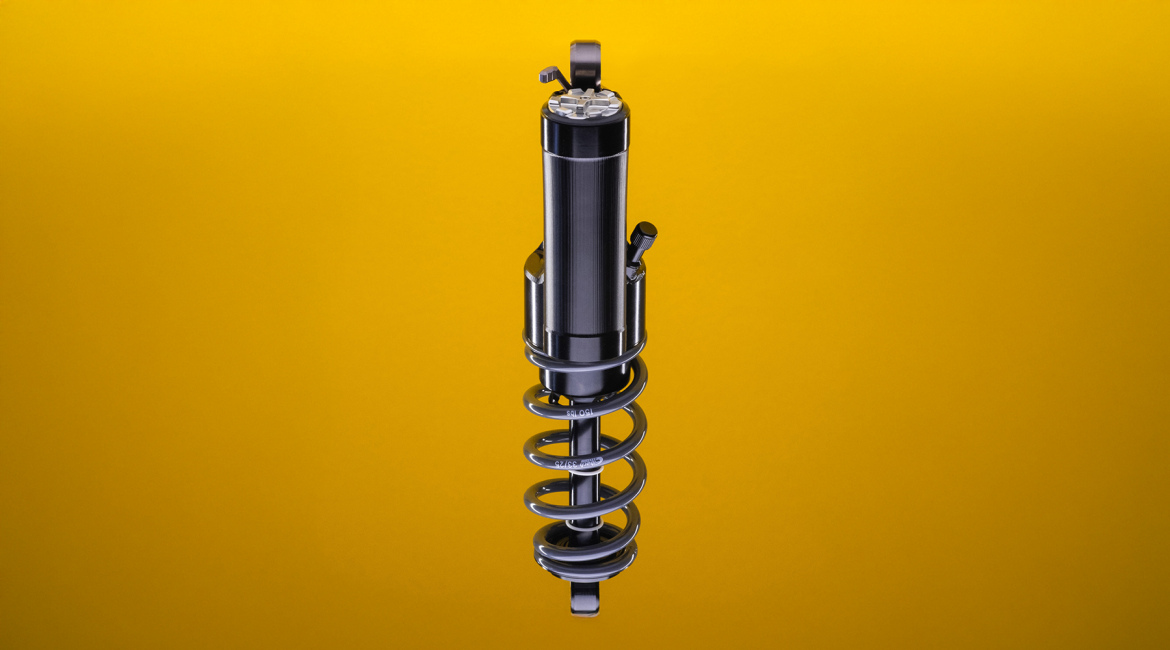 A close-up image of a vehicle shock absorber with a metal coil spring, displayed against a bright yellow background. The shock absorber features a sleek, metallic design and includes adjustment knobs for tuning.