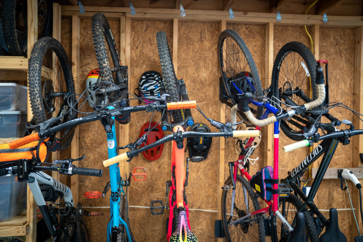 A collection of mountain bikes hanging upside down in a wooden garage. The bikes have various colors and designs, including a turquoise bike, an orange bike, and others with distinct handlebars. Several helmets in different styles are mounted on the wall behind, and there are storage boxes nearby. The scene suggests a well-organized space for cycling enthusiasts.