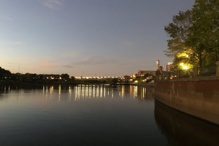 A serene view of a river at dusk, reflecting city lights along the bank. The sky is painted in soft shades of blue and pink, while trees and buildings are illuminated by warm light. The peaceful water surface mirrors the evening ambiance, creating a tranquil scene.