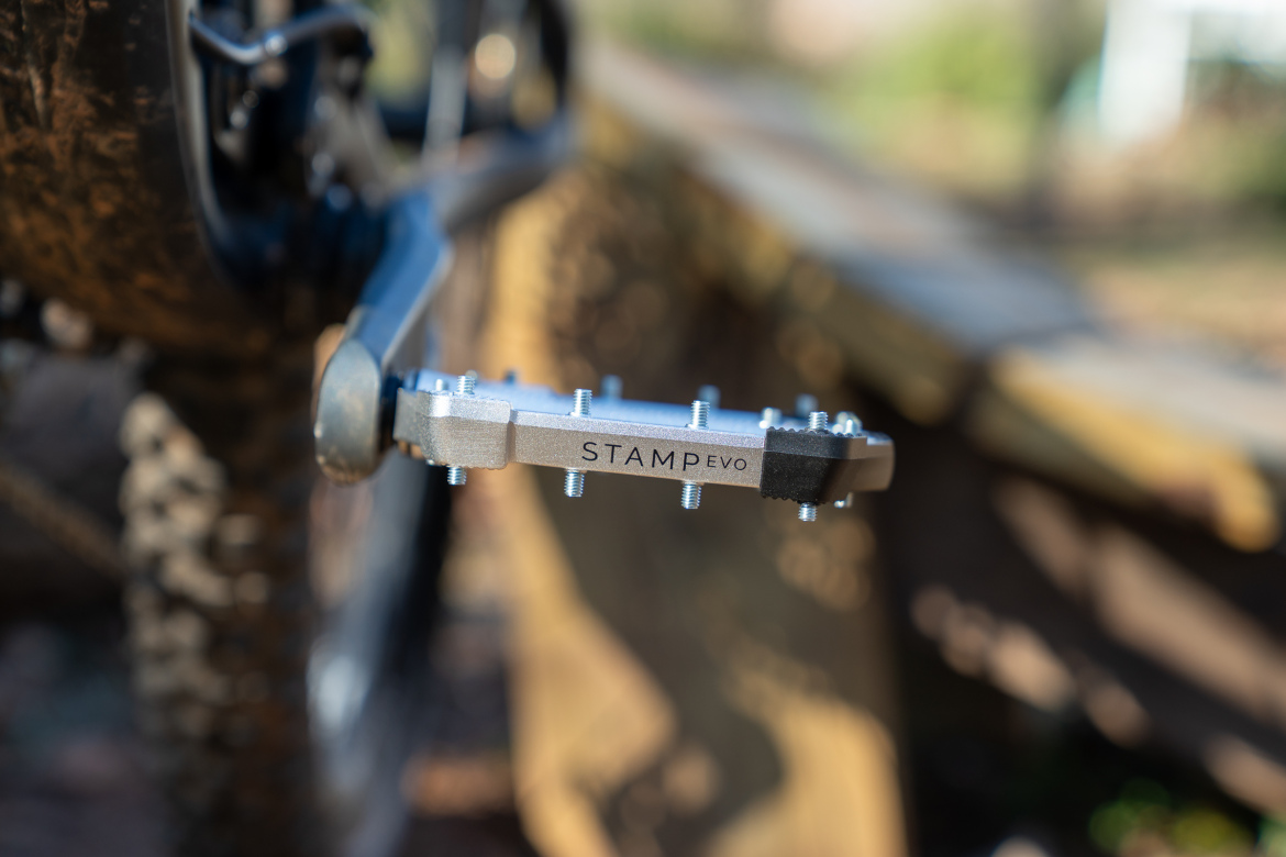Close-up view of a bicycle pedal labeled "STAMP evo," showcasing its metal surface and grip pins, set against a blurred background of a wooden structure in a natural outdoor environment.