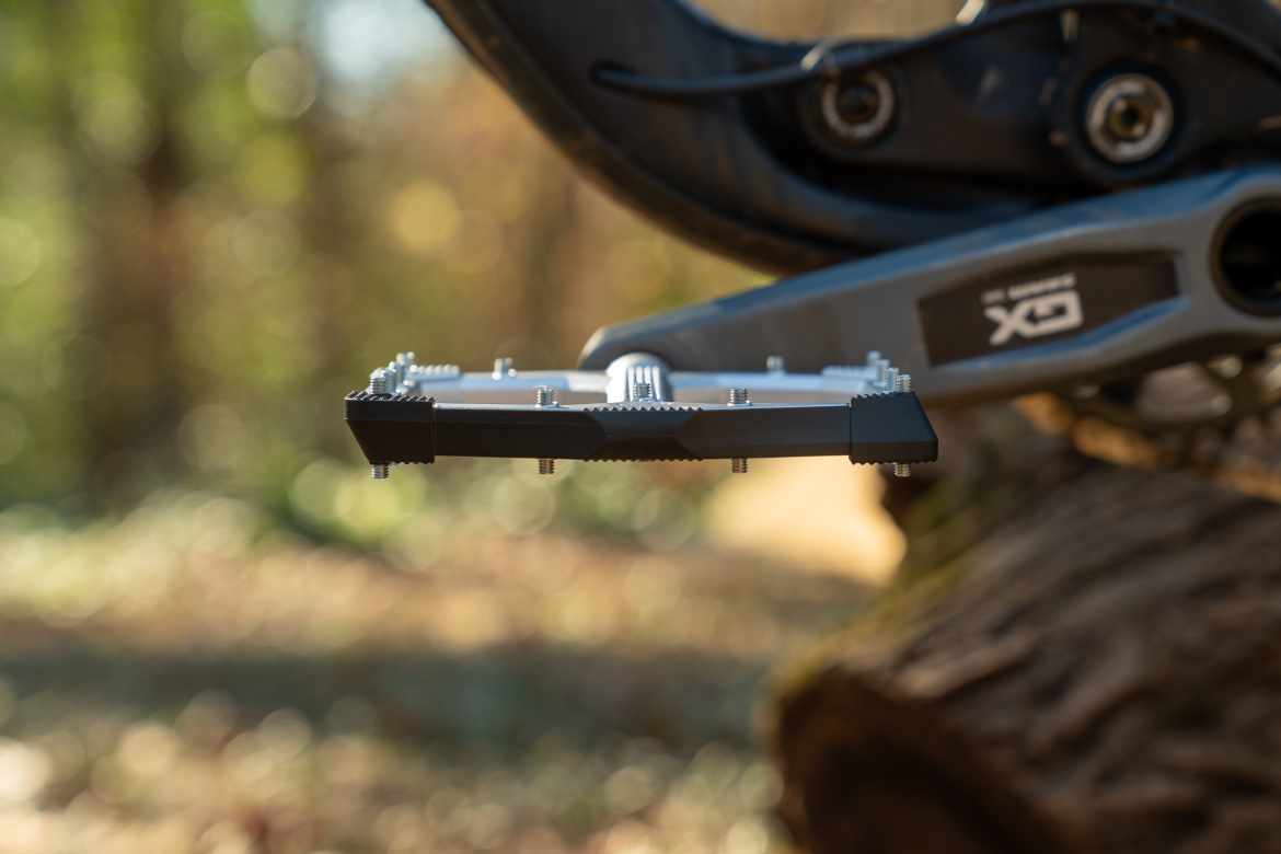 Close-up of a metallic bicycle pedal, featuring a textured surface and spikes for grip, attached to a bike crank in a natural outdoor setting with blurred greenery in the background.