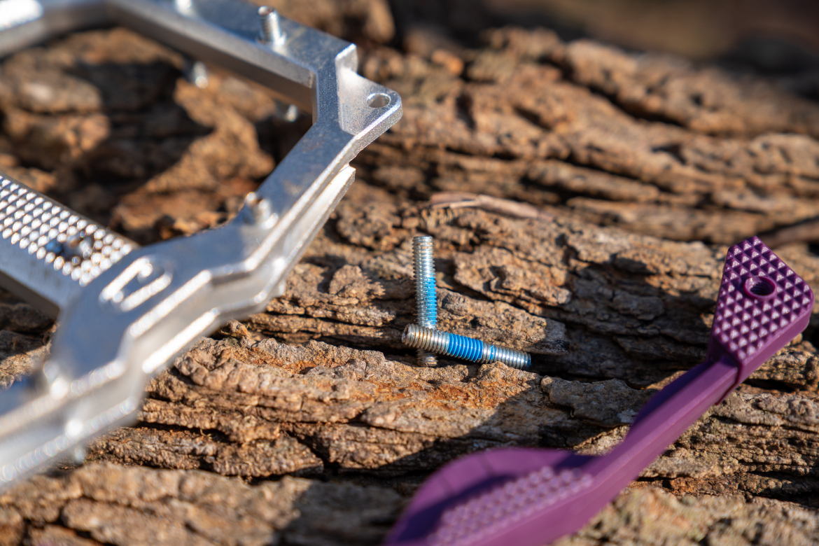 Close-up of a silver metal bicycle pedal, two screws with blue threading, and a purple plastic tool on a textured wooden surface.