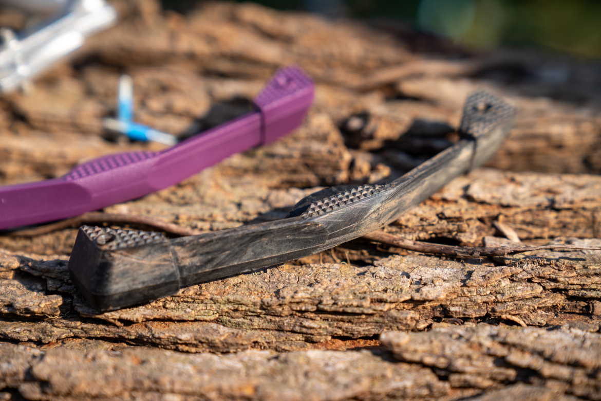 A close-up image of two dental toothbrushes resting on a wooden surface, one black with textured bristles and the other purple, partially out of focus in the background. The wooden surface shows natural texture and color variations.