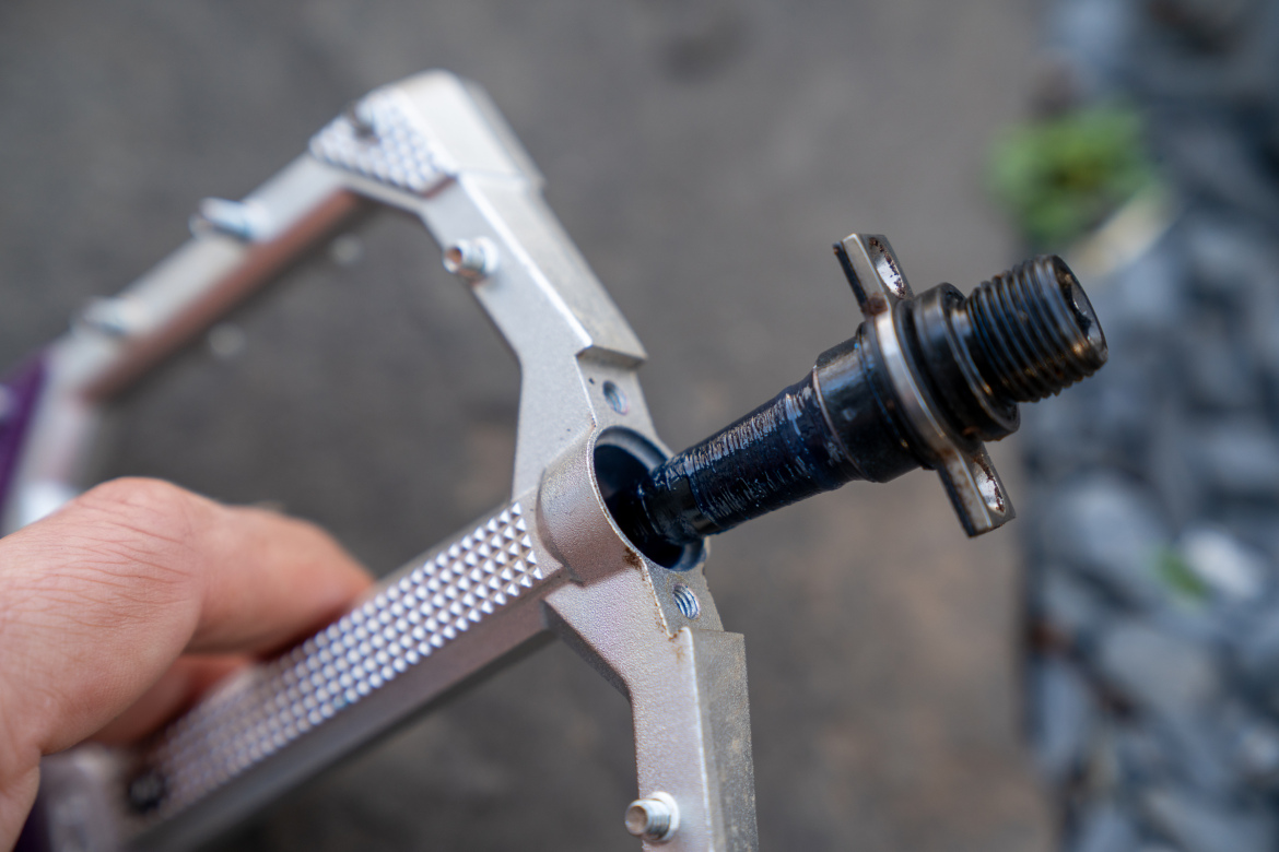 Close-up image of a hand holding a bicycle pedal, showing the metal frame and the threaded spindle. The pedal has a silver finish with textured grip surfaces, while the spindle is dark and partially visible within the pedal's body. The background is blurred, indicating an outdoor setting.