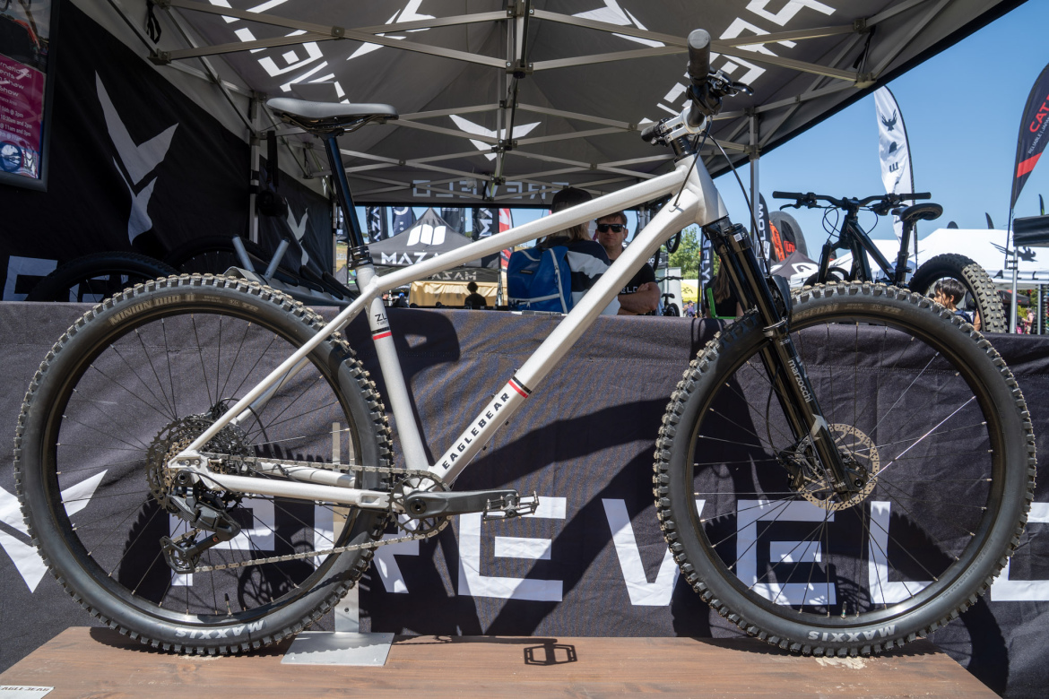 A mountain bike displayed at a tent during an outdoor event, featuring a light-colored frame, thick tires, and a black front fork. The background includes a dark fabric with branding and people engaged in conversation.