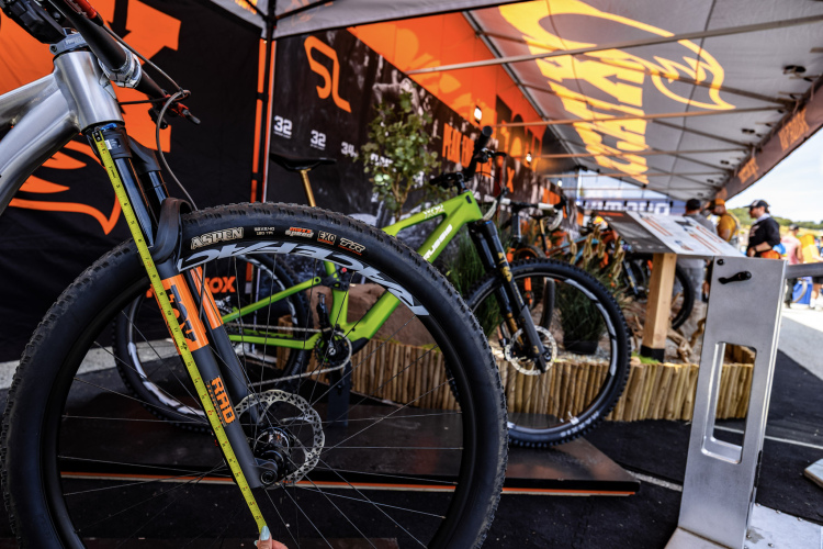 A close-up view of a mountain bike in a display area, featuring a measuring tape alongside the front fork. The background showcases a tent with branding and various bicycles on display. The scene captures the details of the bike's components and an engaging outdoor environment at a cycling event.