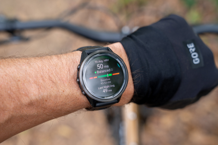 A close-up image of a wrist wearing a smartwatch displaying health metrics, including heart rate variability (HRV) readings. The watch shows a current HRV average of 50 ms and indicates it is balanced, along with baseline values and last night&rsquo;s average. The wrist is partially covered by a black glove, with a blurred background of outdoor scenery.