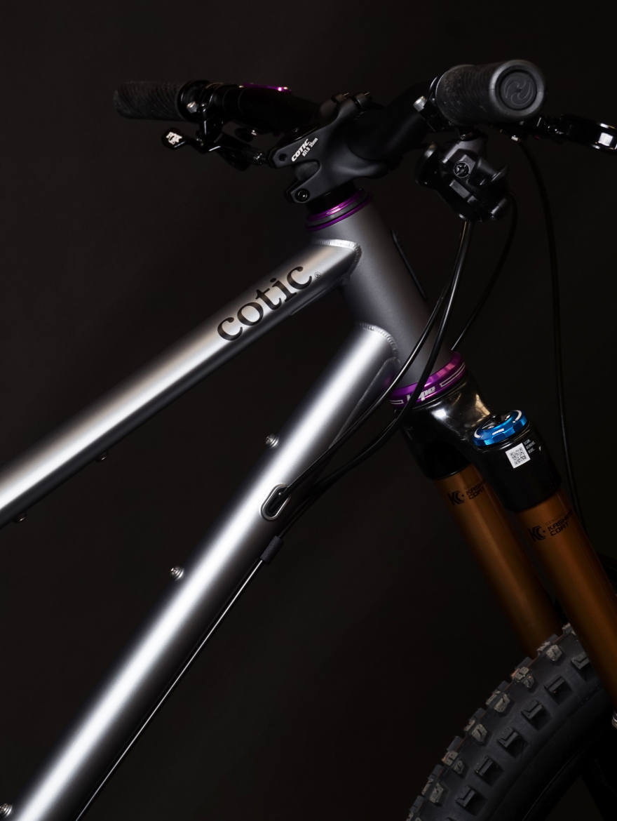 Alt text: A close-up view of a Cotic mountain bike featuring a sleek silver frame with purple accents, handlebars, and front suspension forks. The background is dark, highlighting the bike's details.