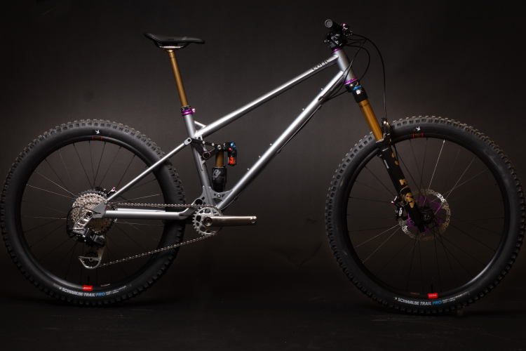 A sleek mountain bike with a aluminum frame featuring a silver finish, equipped with large, rugged tires. The bike has a front suspension fork with golden stanchions, a prominent purple accent on the frame, and a modern drivetrain. The setup emphasizes both performance and aesthetics, with its components arranged in an ideal riding position against a dark background.