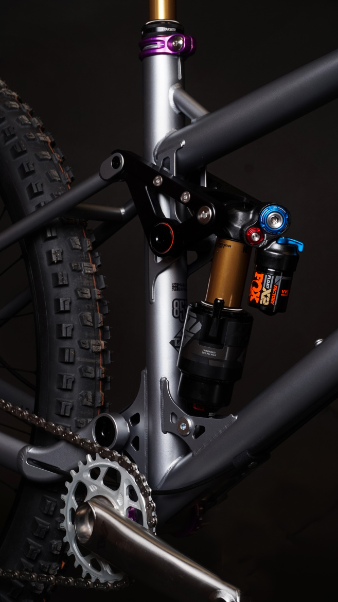 Close-up view of a mountain bike's rear suspension and drivetrain, featuring a gray frame with a visible shock absorber, detailed linkage, and a chainring. The image highlights components like the gold and purple shock mounts, along with the rear tire and chain. The background is dark, emphasizing the bike's design.