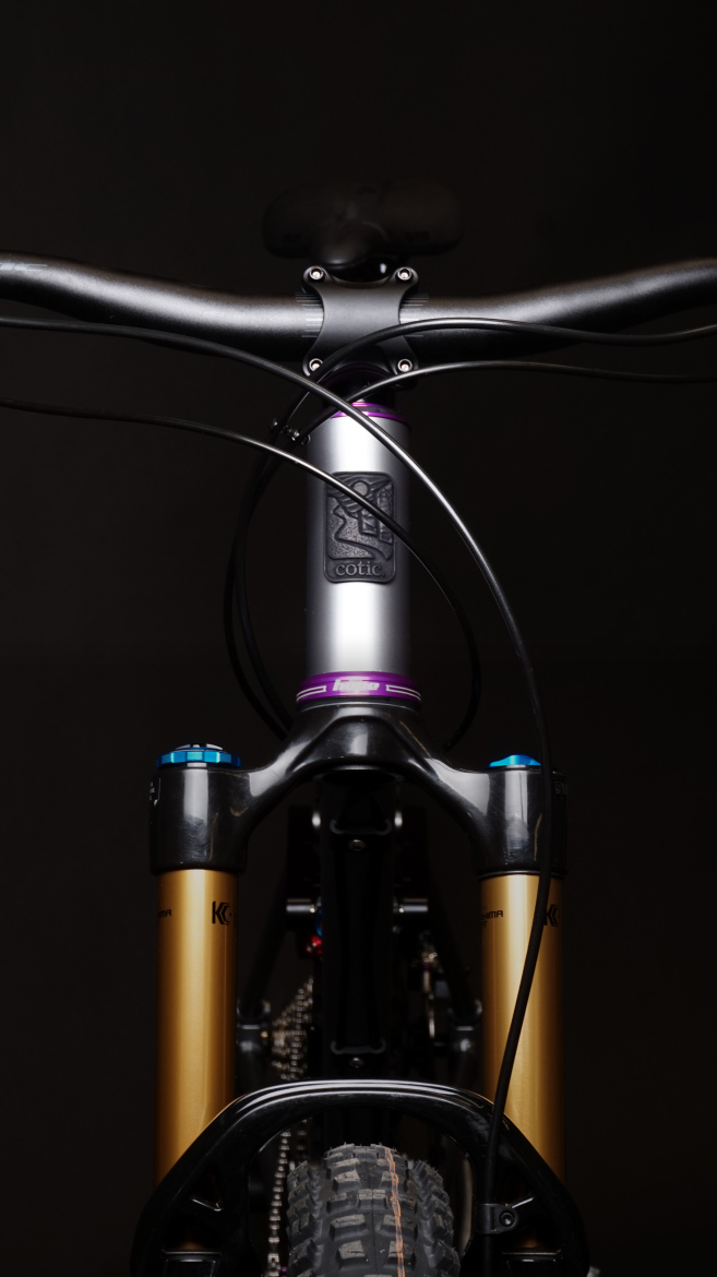 Close-up image of a mountain bike from the front, showcasing the handlebars, stem, and front fork. The frame is dark, with prominent gold suspension components and a silver stem featuring the brand logo. Cables run neatly along the frame, and the background is black, highlighting the bike's details.