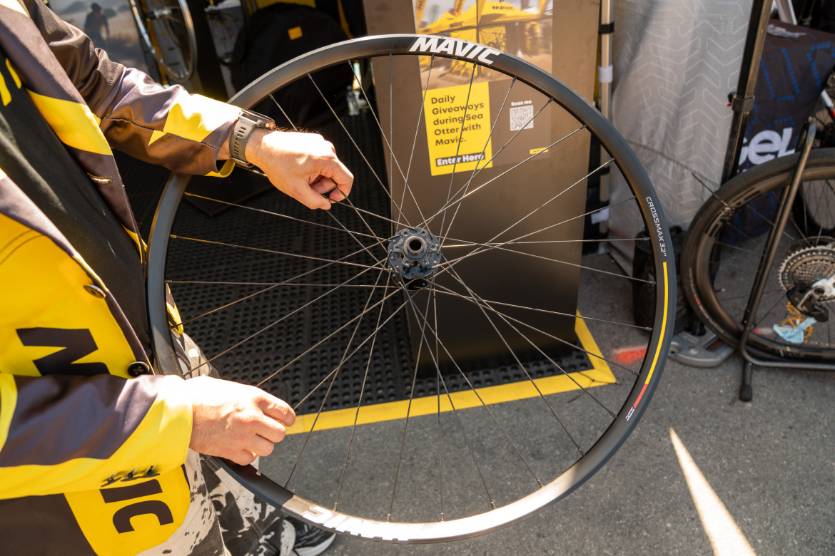 A person wearing a yellow and black jacket is holding a bicycle wheel with visible spokes and a hub, set against a backdrop featuring promotional materials for Mavic. The scene is outdoors, possibly at an event like a bike festival.