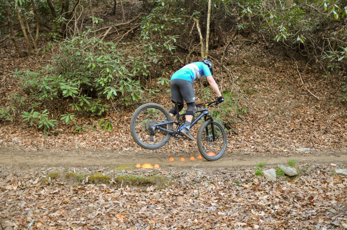 A mountain biker performing a trick on a dirt trail surrounded by greenery and autumn leaves, with orange cones set up on the ground.