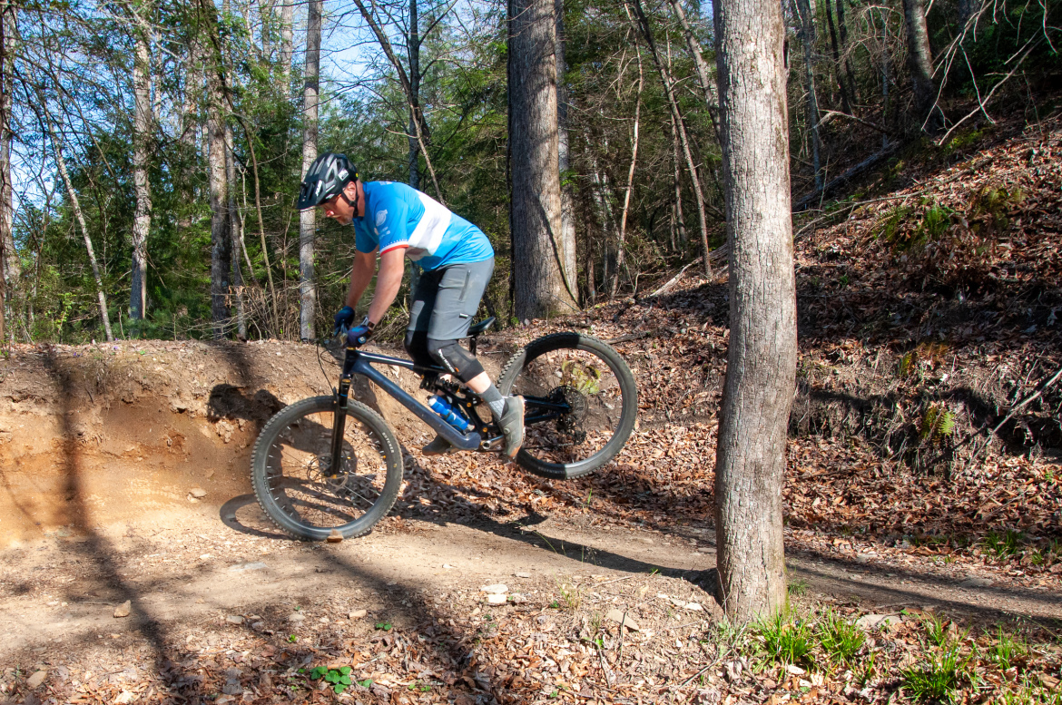 A mountain biker in a blue and white jersey performs a jump on a dirt trail surrounded by trees and autumn leaves. The rider is wearing a helmet and protective gear, focusing on the jump over a small ditch. The background features a wooded area, capturing the essence of outdoor cycling.