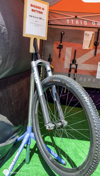 A bicycle fork displayed at a trade show, labeled with a sign that reads "BIGGER IS BETTER! E36 Fork for 32-inch wheel." The fork is attached to a wheel with a textured tire, sitting on a green surface inside a tent. Various other bike components are visible in the background.