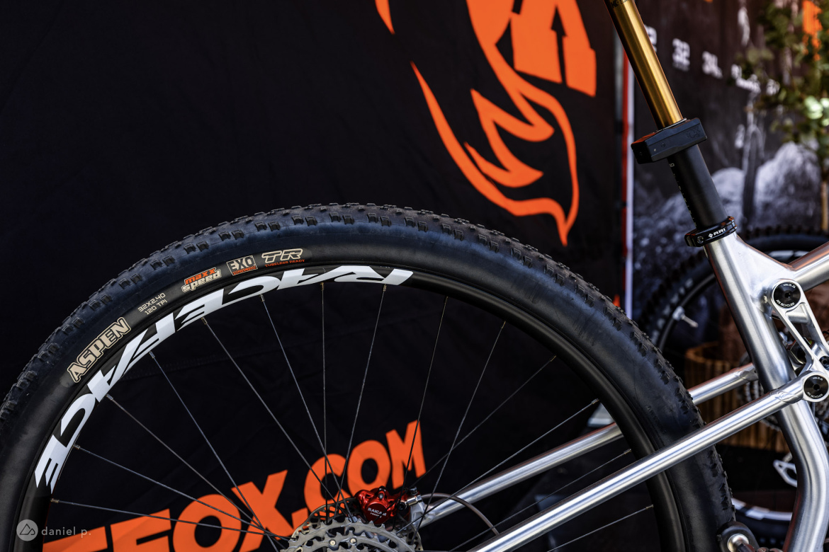 A close-up view of a bicycle wheel featuring a distinctive tire with the brand name "Aspen" visible. The wheel is attached to a sleek silver bike frame, with a red brake component visible. In the background, a black banner with an orange graphic can be seen, suggesting a bike or outdoor brand. The image captures the details of bicycle components, showcasing craftsmanship and design.