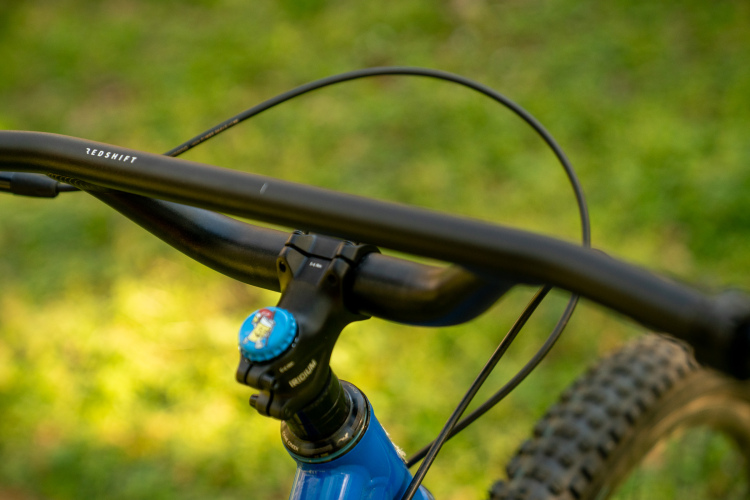 Close-up view of a mountain bike's handlebars, featuring a black handlebar and stem, with a blue adjustment knob visible. The background shows a blurred green grassy surface, suggesting an outdoor setting.