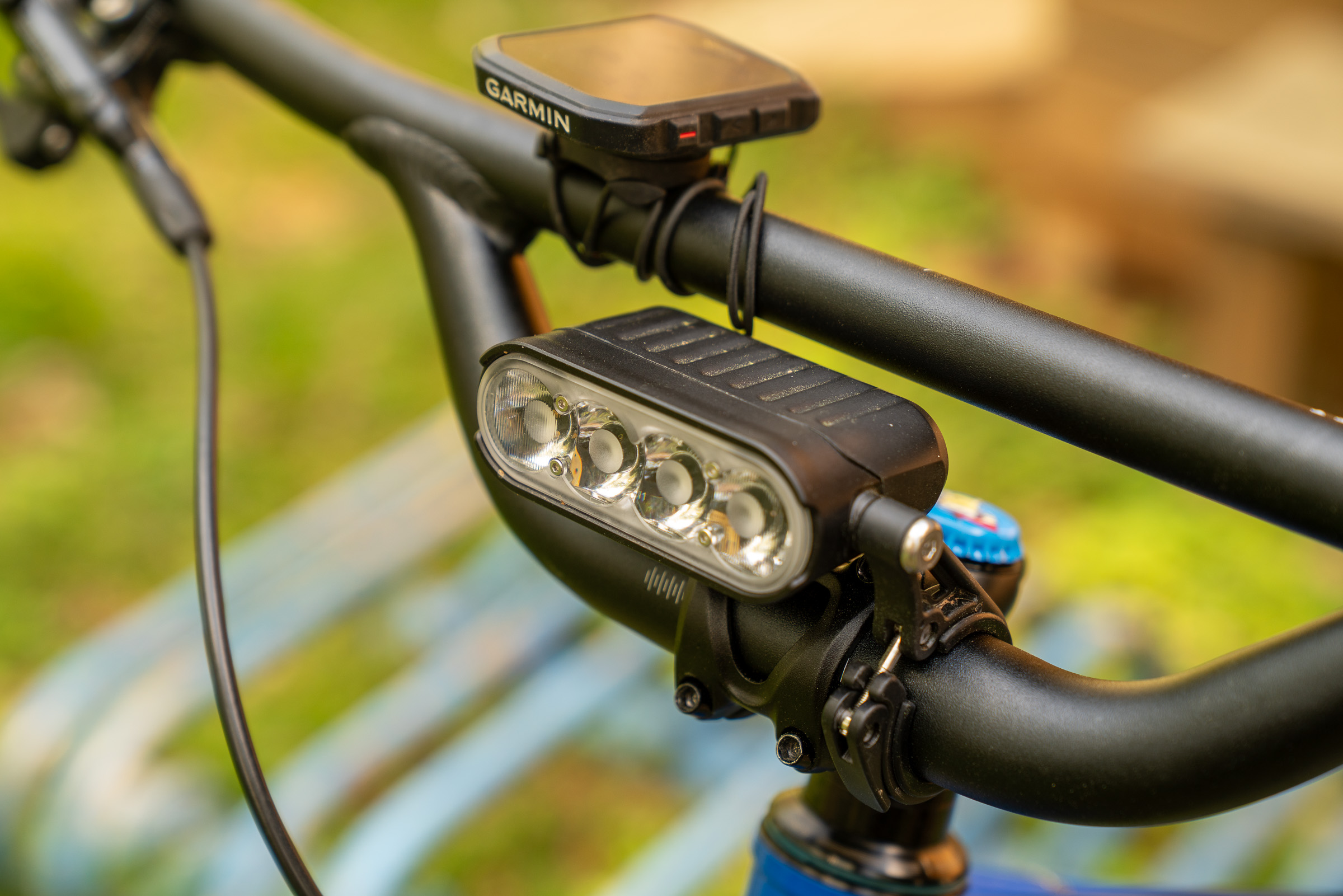 Close-up of a bicycle handlebar featuring a mounted bike light with multiple LED bulbs and a Garmin device for navigation or tracking. The background is blurred with green grass and blue bicycle frame elements visible.