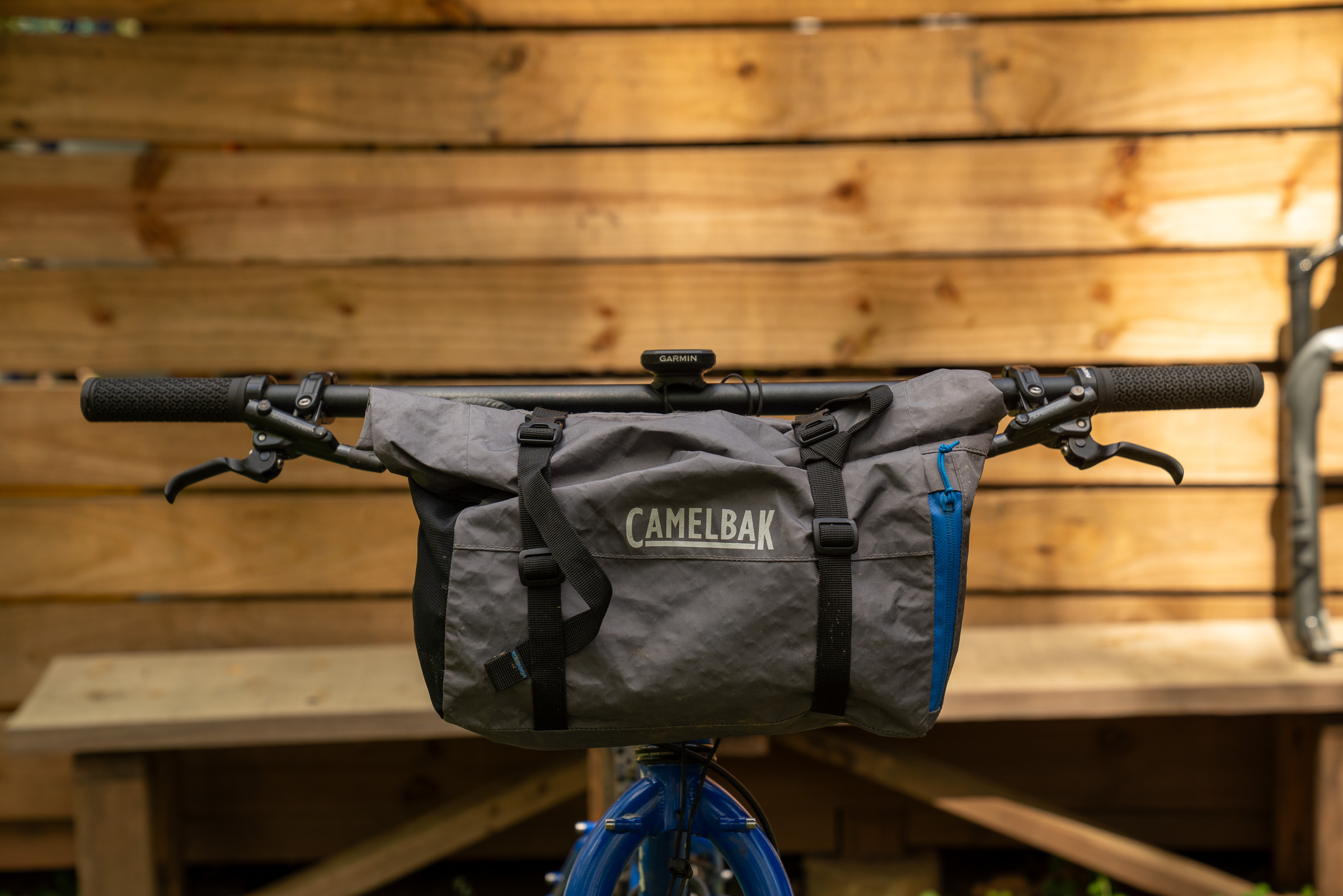 A close-up view of a bicycle handlebar featuring a gray CamelBak bag secured to the front. The bag has blue accents and is fastened with adjustable straps. In the background, there's a wooden wall with a natural finish, complementing the outdoor setting.
