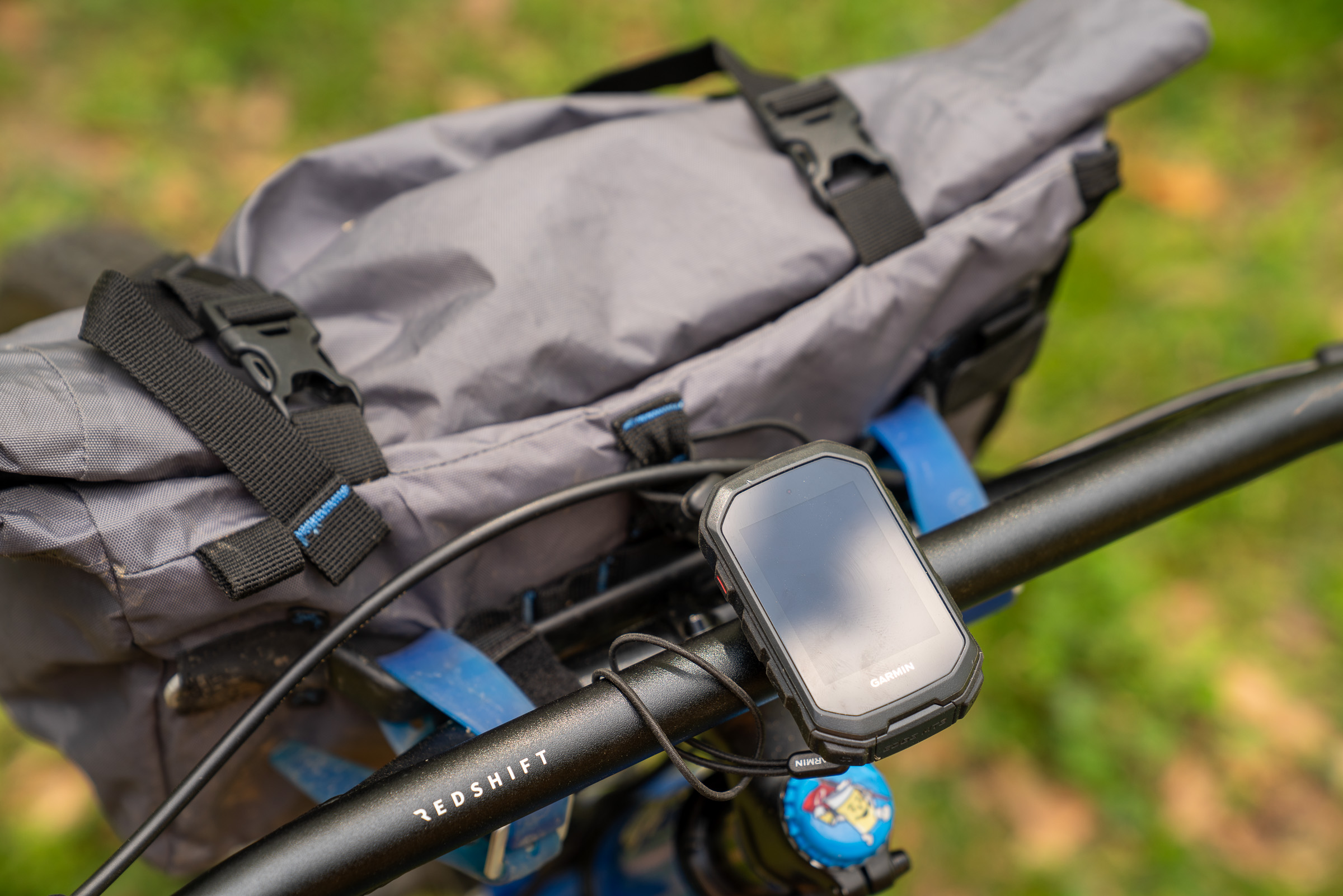 A close-up view of a bicycle handlebar featuring a mounted GPS device and a backpack. The GPS is connected by a cable, and the backpack, made of gray fabric, has clipped straps. The handlebar is marked with the word "REDSHIFT," and a colorful object can be seen at the end of the handlebar. The background is blurred with hints of greenery.