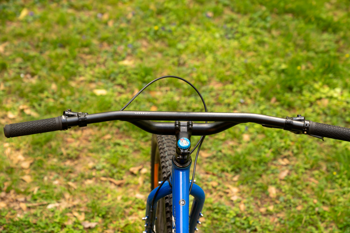 Close-up view of mountain bike handlebars with black grips, positioned over the bike's blue frame. The background features green grass and scattered leaves.