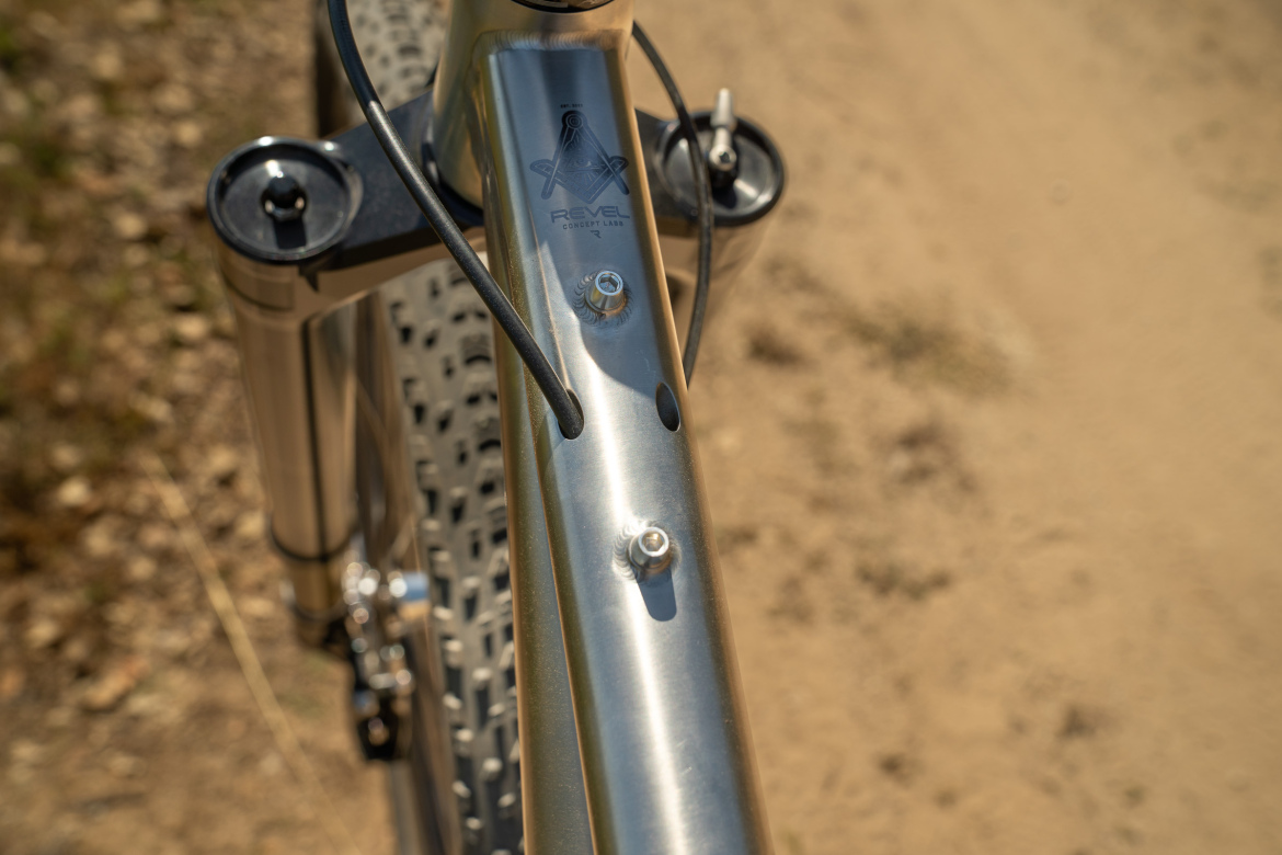 Close-up view of a bicycle frame, highlighting the shiny metallic top tube with the Revel logo, along with mounting points and cables. The background features a dirt path with scattered rocks and grass.