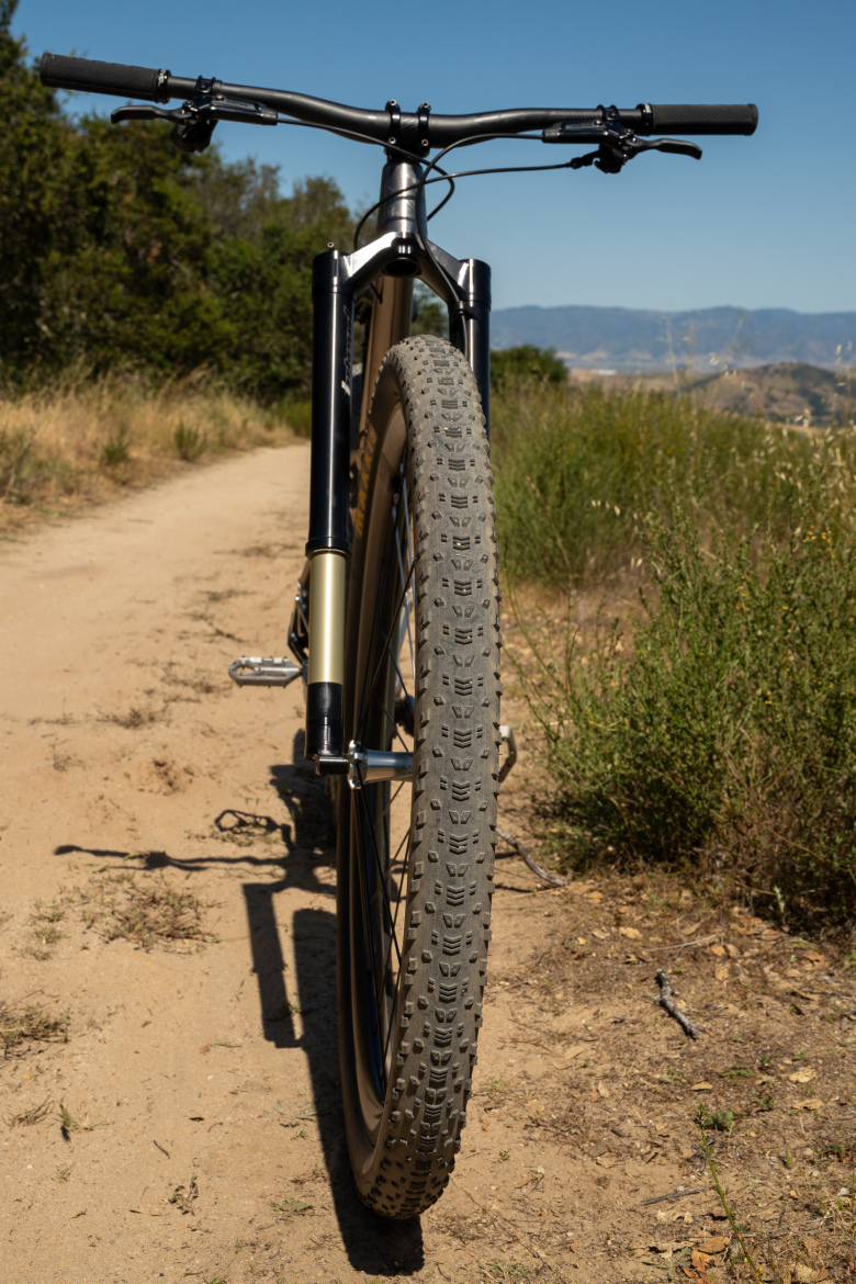 A close-up view of a mountain bike in a natural outdoor setting, facing down a dirt trail. The bike's front wheel, tire, and handlebars are prominently featured, with foliage and rolling hills visible in the background under a clear blue sky.