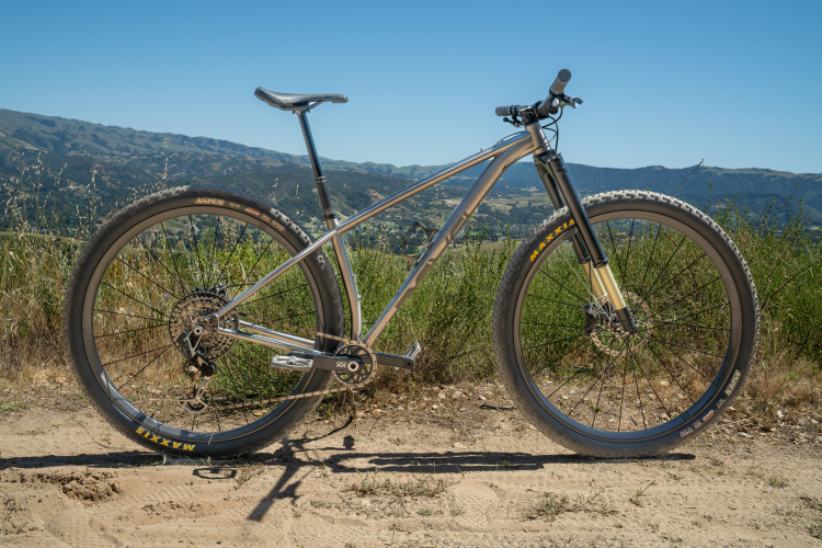 A mountain bike with a sleek silver frame stands on a dirt path surrounded by greenery, with rolling hills in the background under a clear blue sky. The bike features wide tires and a front suspension fork, showcasing its readiness for off-road trails.