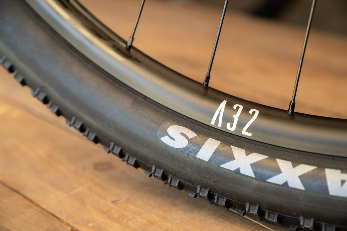 Close-up of a mountain bike tire and wheel rim, showcasing the tread pattern and markings. The rim is labeled "A32," and the tire features the name "SIXAX." The background is a soft, blurred wooden surface.