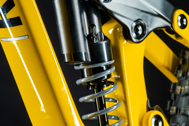 Close-up of a yellow bicycle frame showcasing the rear shock absorber and spring mechanism. The image highlights the black suspension parts and metallic components, emphasizing the engineering and design of the bike's suspension system against a dark background.