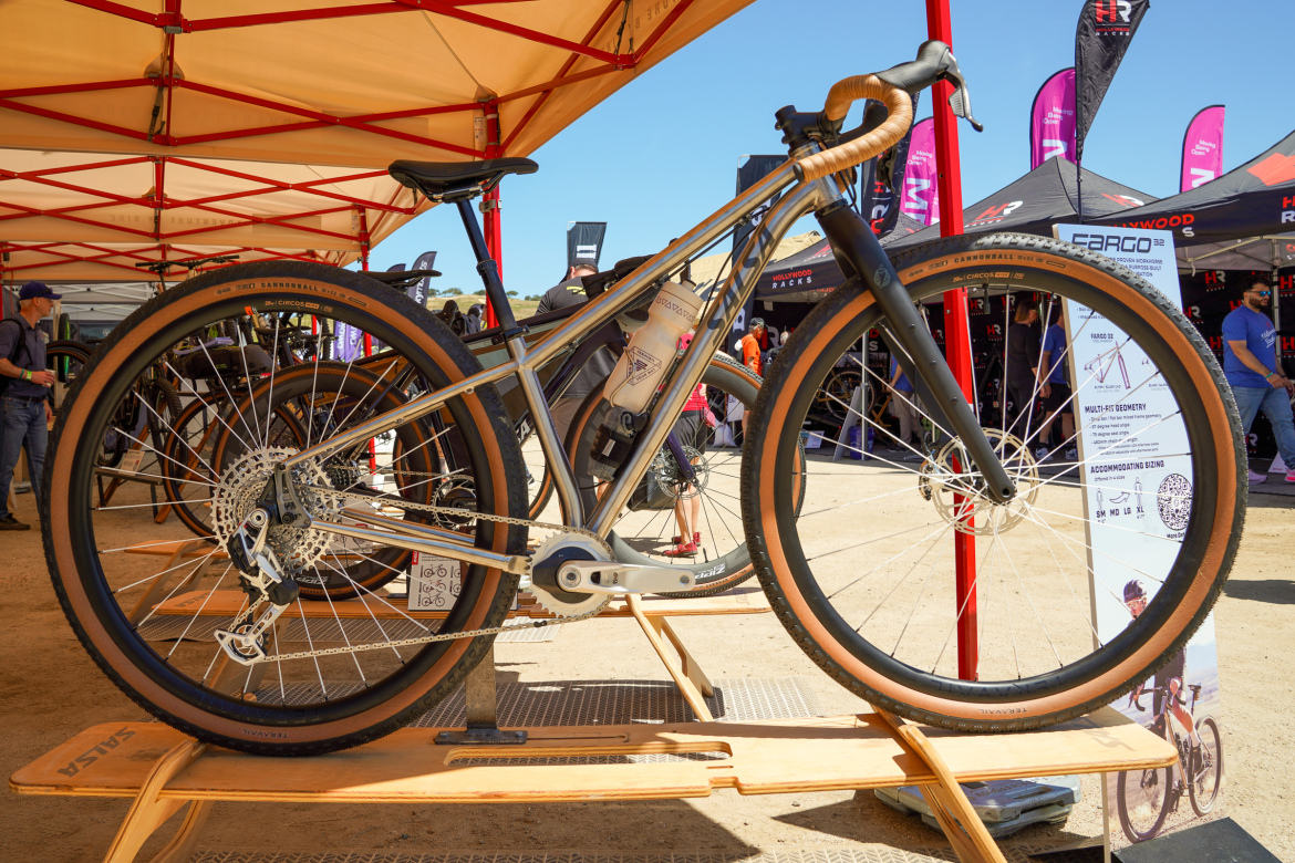 A modern bicycle displayed at a cycling event, featuring a sleek aluminum frame, wide tires with tan sidewalls, and a battery mounted on the frame. The bike is positioned on a wooden rack with red and yellow tents in the background, showcasing various bike brands. Sunlight is casting a bright light on the scene, highlighting the bike's details.