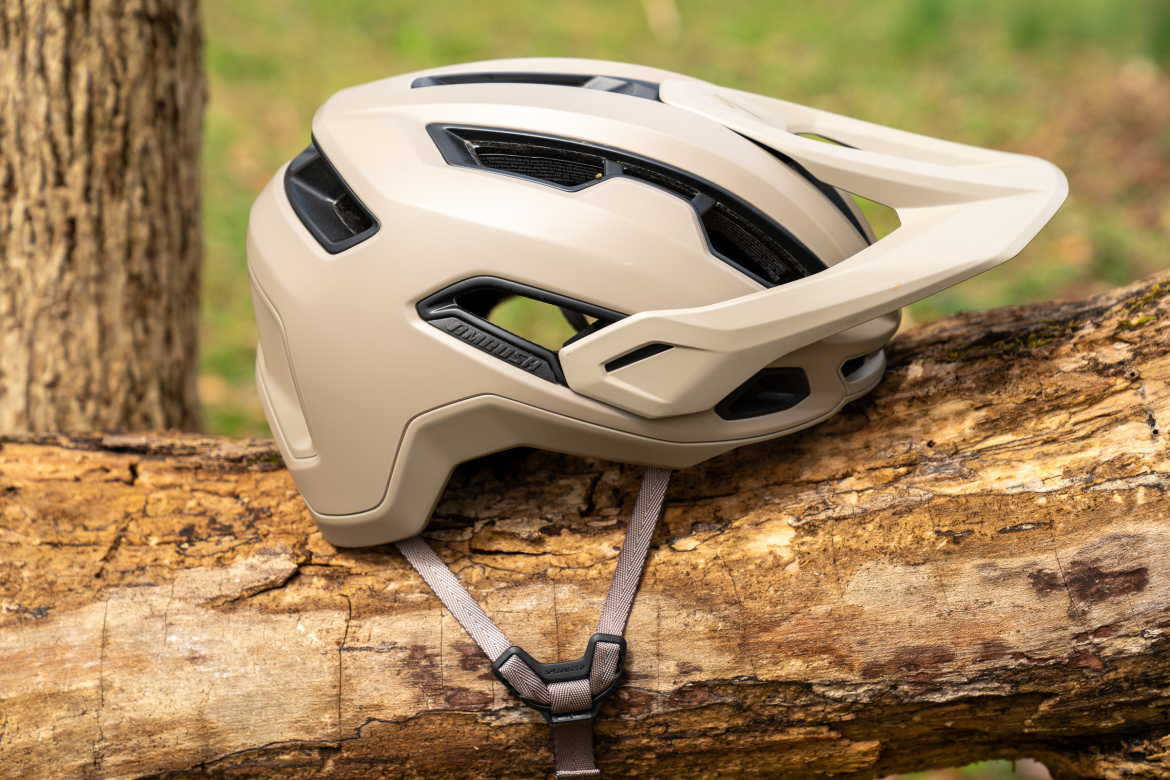 A beige bicycle helmet resting on a log, showcasing its aerodynamic design and ventilation features. The helmet is positioned on its side, highlighting the straps and inner padding against a natural background of greenery.