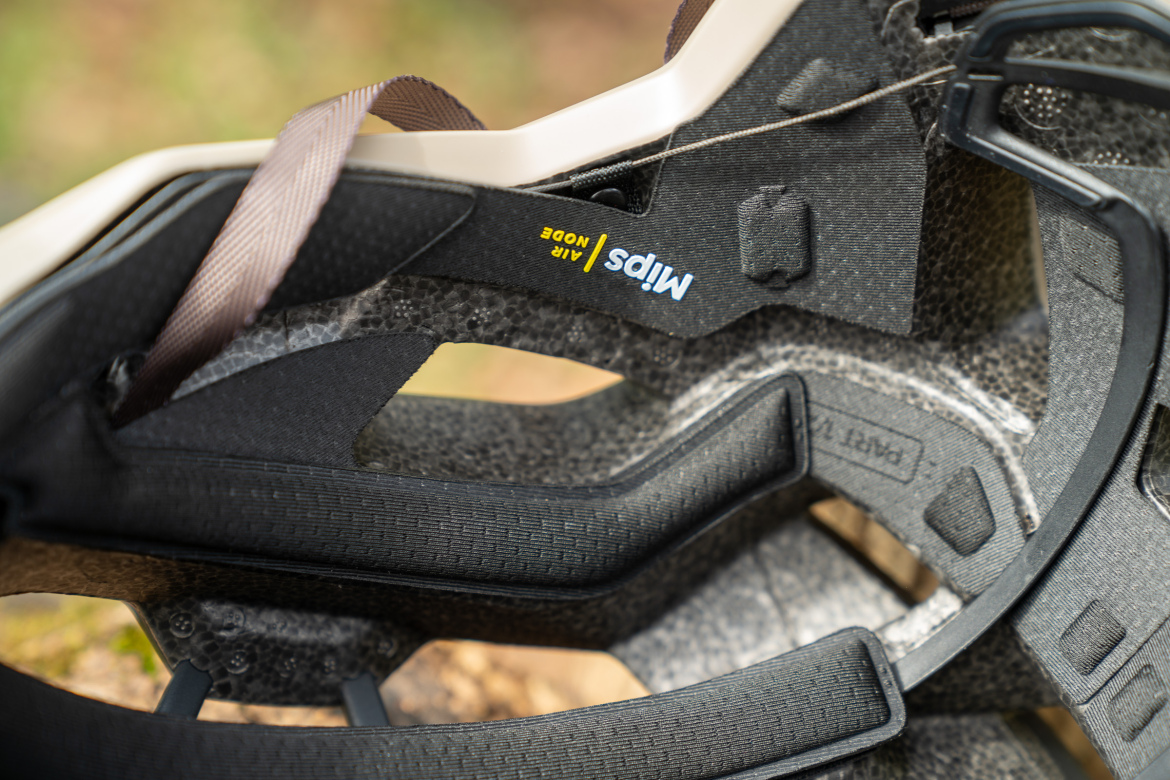 Close-up image of the interior of a cycling helmet, showcasing the padding and adjustable straps. The inner structure features ventilation holes and textured materials, with a label indicating "MIPS AIR NODE" for enhanced safety technology. The background is blurred, emphasizing the details of the helmet's design.