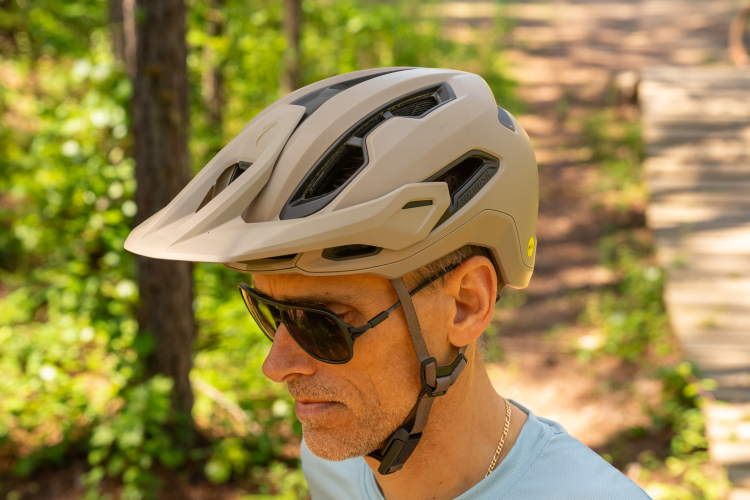 A side profile of a man wearing a light-colored mountain biking helmet and dark sunglasses, standing on a wooded path surrounded by greenery.