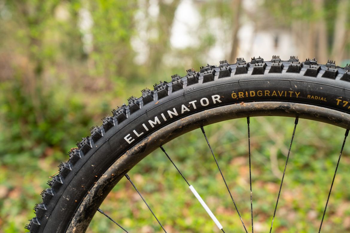 Close-up view of a mountain bike tire labeled "Eliminator" with a rugged tread pattern, showcasing its durability. The tire is set against a blurred natural background with greenery.