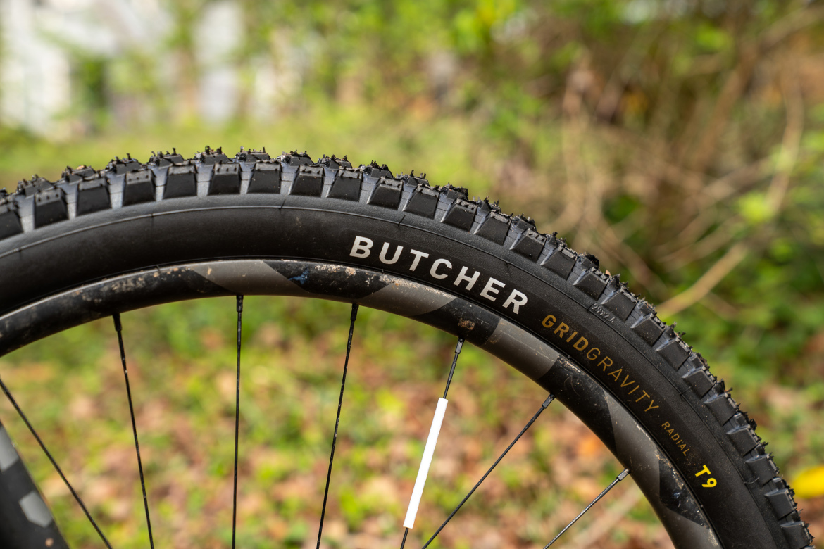 Close-up of a mountain bike tire featuring the brand name "Butcher" and the model "GridGravity T9." The tire has a rugged tread pattern designed for off-road traction, set against a blurred green background of foliage.