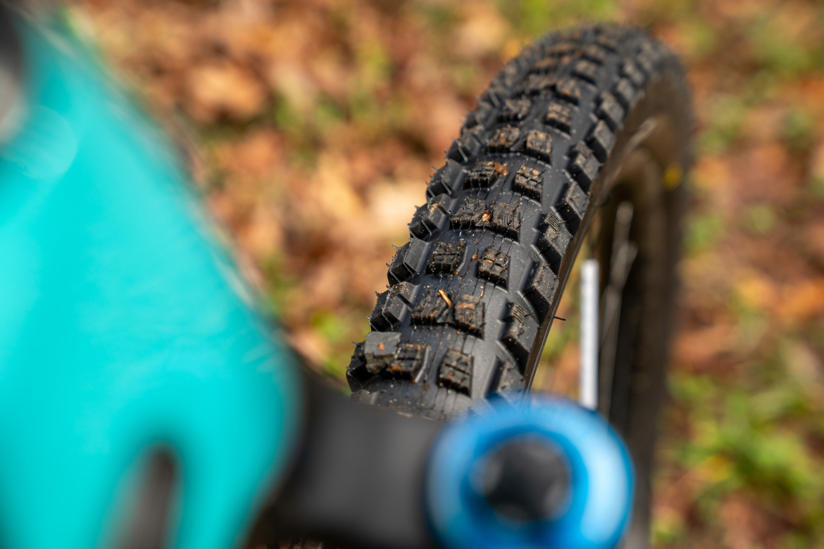 Close-up view of a mountain bike tire with a rugged tread pattern, partially covered in dirt and debris, set against a blurred background of autumn leaves. The image also features a turquoise bike frame and a blue component.
