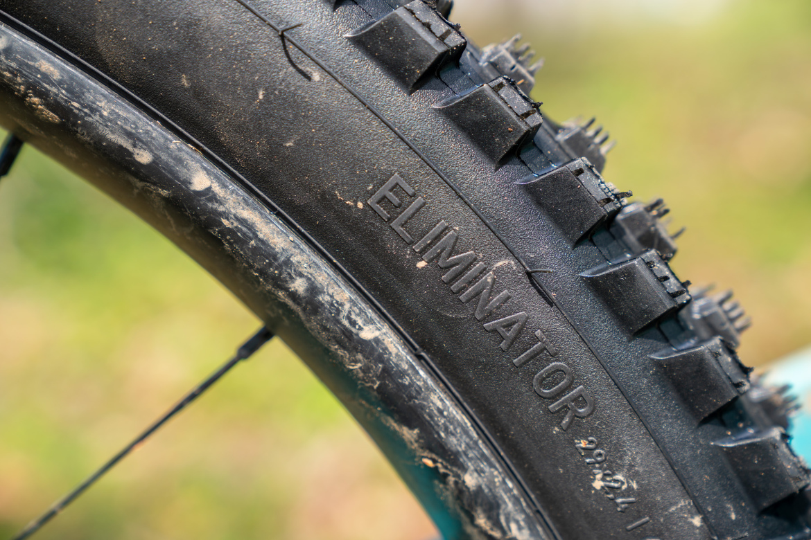 Close-up of a mountain bike tire with the word "ELIMINATOR" embossed on the sidewall, featuring a rugged tread pattern and a layer of dust along the rim. The background is blurred, highlighting the tire.