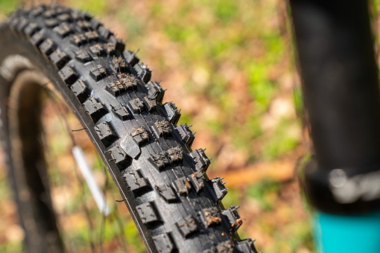 Close-up view of a mountain bike tire, showing its rugged tread pattern and texture, with dirt and debris visible on the surface. The background features a blurred natural setting with green grass and blurred foliage.