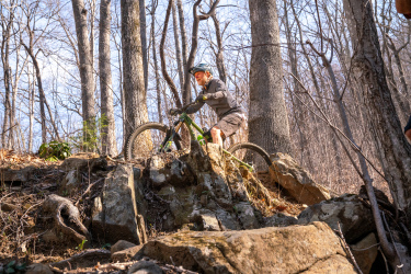 A mountain biker navigating rocky terrain in a forested area, surrounded by tall trees and bare branches. The rider is wearing a helmet and is positioned on a rock ledge, focusing on the path ahead. Sunlight filters through the trees, illuminating the landscape. Spencer Branch mountain bike trail.