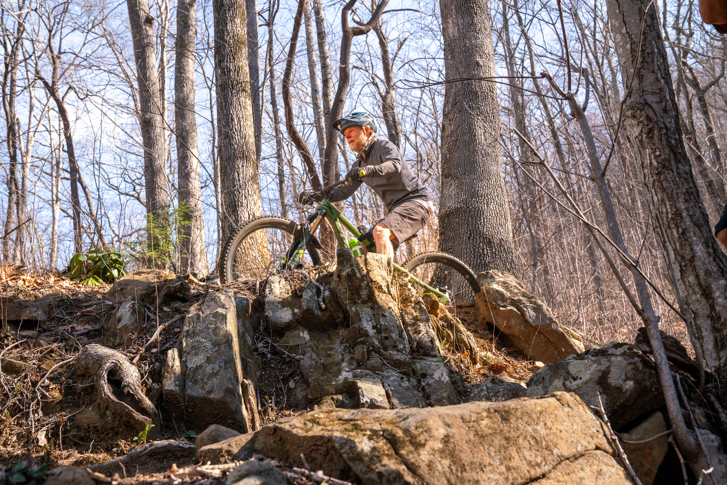 A mountain biker navigating rocky terrain in a forested area, surrounded by tall trees and bare branches. The rider is wearing a helmet and is positioned on a rock ledge, focusing on the path ahead. Sunlight filters through the trees, illuminating the landscape. Spencer Branch mountain bike trail.