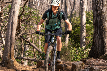 A mountain biker navigating a rocky trail in a forested area, wearing a helmet and riding gear. The scene features bare trees and greenery in the background, indicating an early spring setting. The biker is smiling and focused on maneuvering over the rocks. Spencer Branch mountain bike trail.