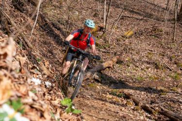A person riding a mountain bike on a narrow, dirt trail in a wooded area, surrounded by fallen leaves and small wildflowers. The rider is wearing a red and black jersey, black shorts, and a light blue helmet, navigating past a fallen log on the trail. The scene captures the essence of outdoor adventure and mountain biking.