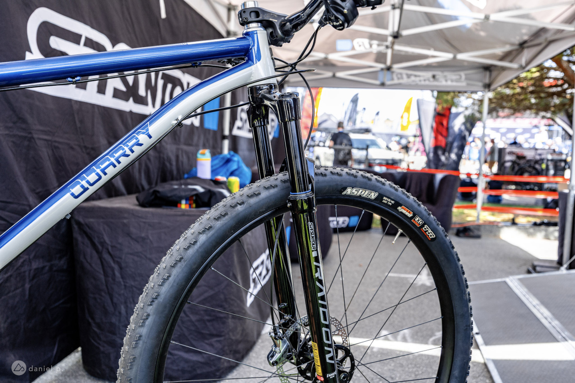 A close-up view of a blue mountain bike with a prominent "Quarry" label on the frame. The image showcases the front fork, wheel, and disc brake, set against a background of a vendor tent and various biking equipment on display.