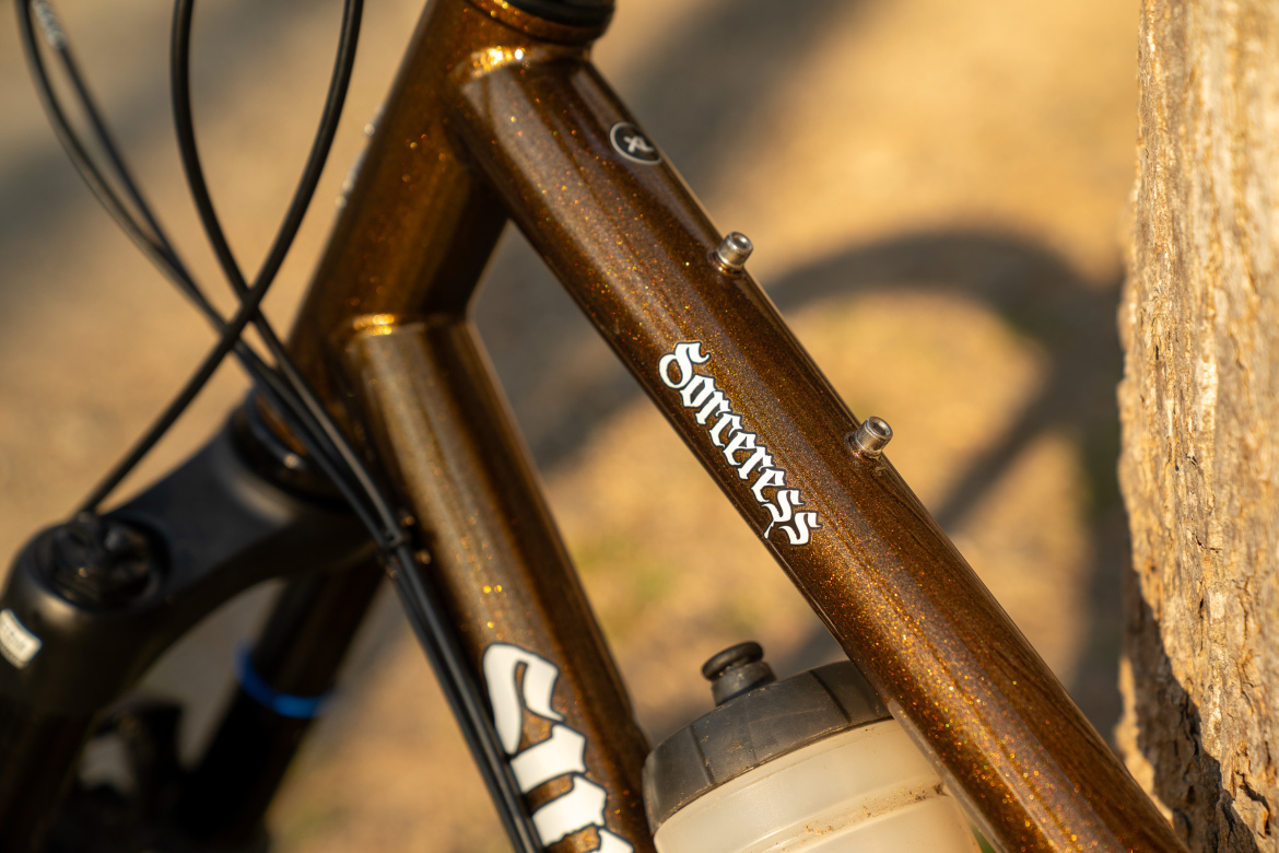 Close-up of a bike frame with a sparkling brown finish, featuring the word "Sorceress" in white lettering. A black water bottle holder is attached, and part of the bike's suspension system is visible. The background consists of a blurred natural setting.