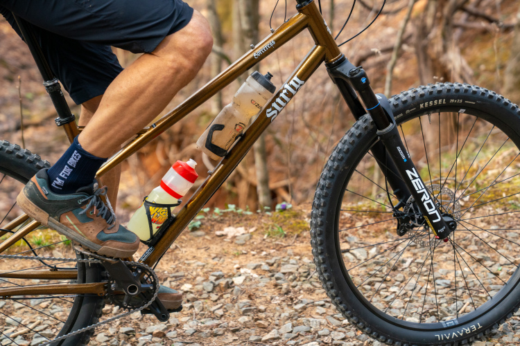 A close-up view of a cyclist's leg and foot on a mountain bike, showcasing a brown bike frame with a water bottle holder attached. The cyclist wears dark shorts and a pair of colorful socks. The bike features a thick front tire and a visible chainring, with gravel and foliage in the background.