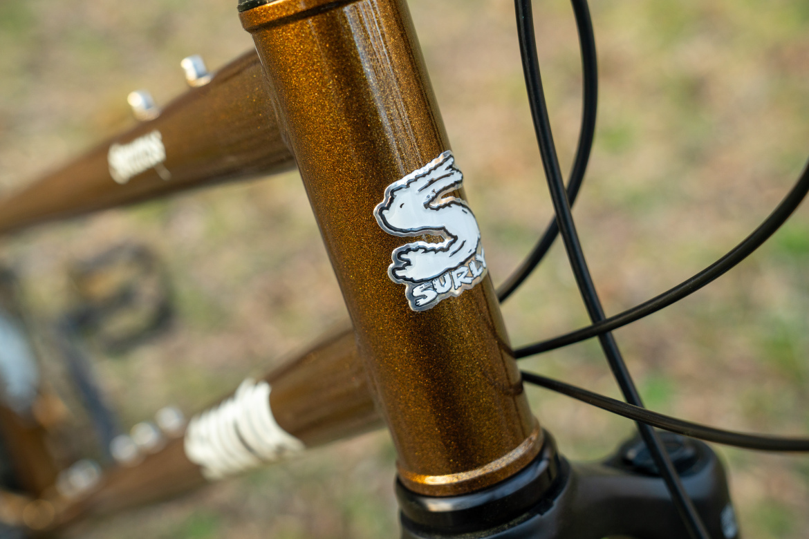 Close-up of a bicycle's headtube featuring a shiny brown metallic finish and a Surly logo sticker shaped like a rabbit. The background shows a blurred natural setting.
