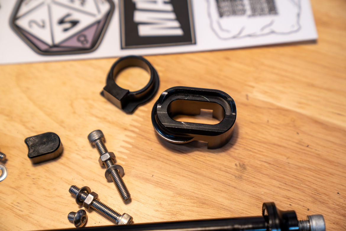 Close-up of various mechanical components on a wooden surface, including a black plastic piece with an oval opening, a circular black ring, a small black cap, and several metal screws and washers, along with printed materials in the background featuring gaming elements.