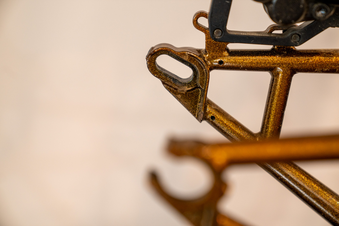 Close-up view of a metallic bicycle frame showing intricate details and a glossy, gold finish. The image captures the frame's connection points and structural design against a soft, blurred background.
