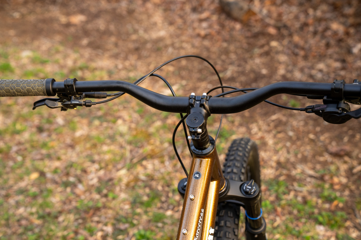 A close-up view of a mountain bike's handlebars, featuring textured grips and gear shifters. The bike's frame is a shiny gold color, and the background shows an outdoor setting with brown foliage and greenery.